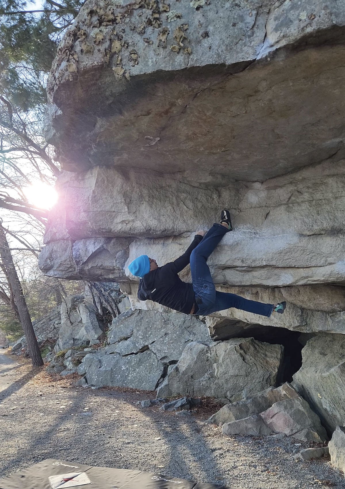 A climber wearing a blue hat and black jacket is bouldering on an overhanging rock formation outdoors, with the sun shining through the trees in the background