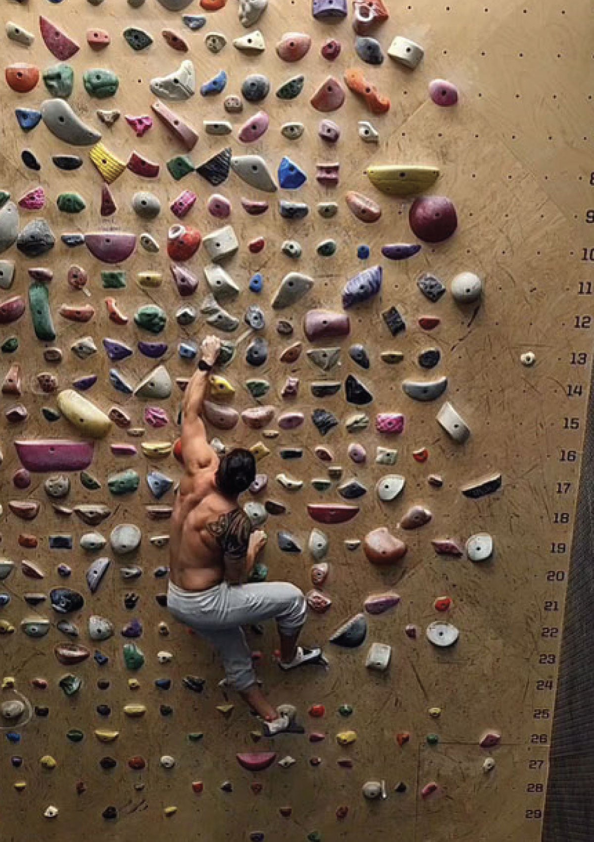 A shirtless climber with a tattoo on his shoulder is bouldering on an indoor climbing wall filled with various colorful holds