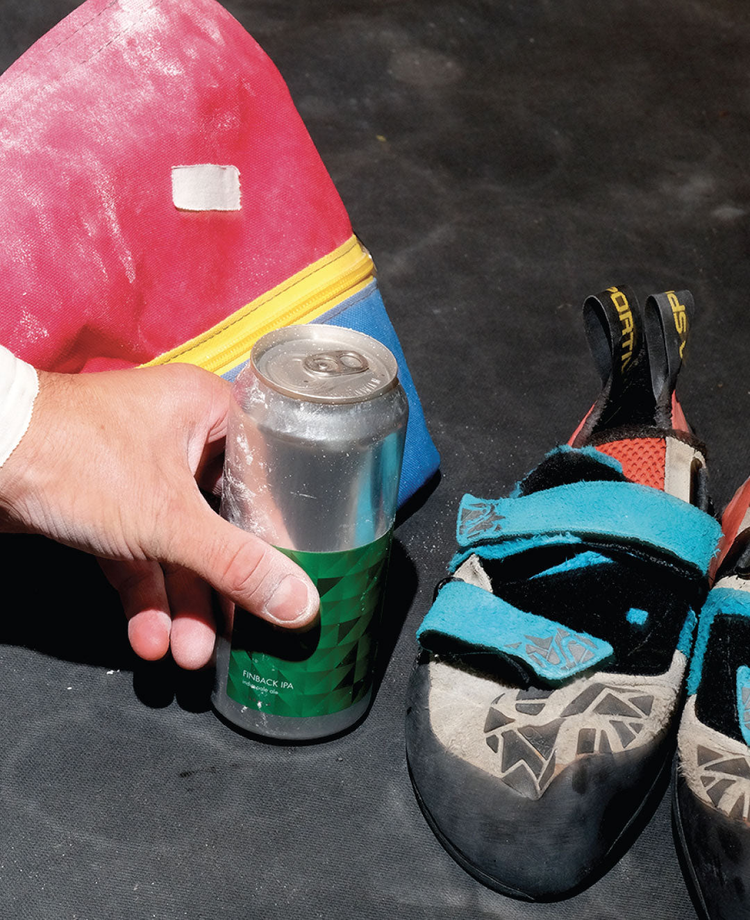 A hand holds a canned beverage next to a pair of well-used climbing shoes and a chalk bag on a black surface